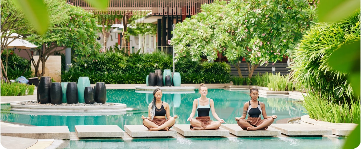 Three women meditating cross-legged beside a resort pool in a tropical garden