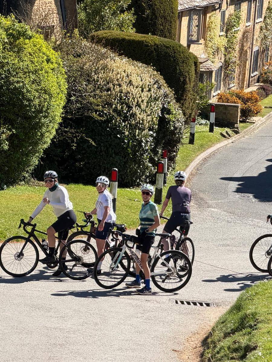 A group of cyclists smiling at the camera on a sunny Cotswolds village lane